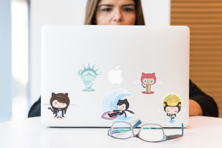 A laptop decorated with colorful stickers including a GitHub Octocat sticker, sitting on a white table with eyeglasses in the foreground